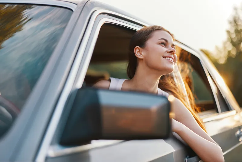 Happy young woman enjoying a road trip in a car with her head out the window, golden hour light, carefree spirit, and vibrant nature background.