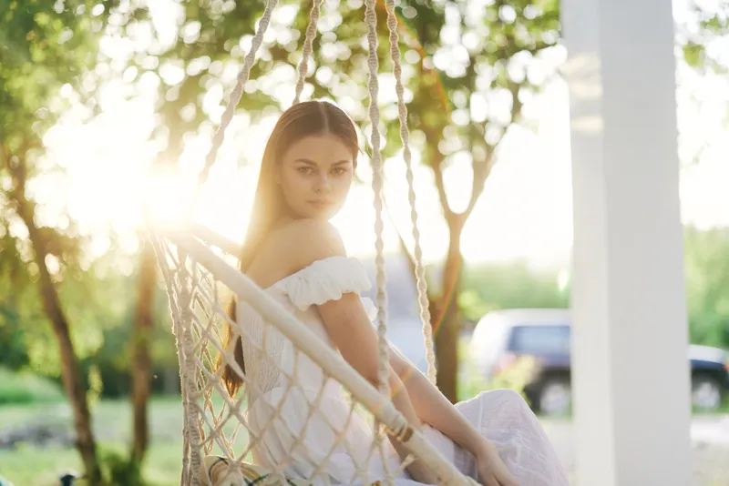 Young woman in white dress sitting in a swing, basking in sunlight, surrounded by greenery, evoking feelings of serenity and nature in summer setting