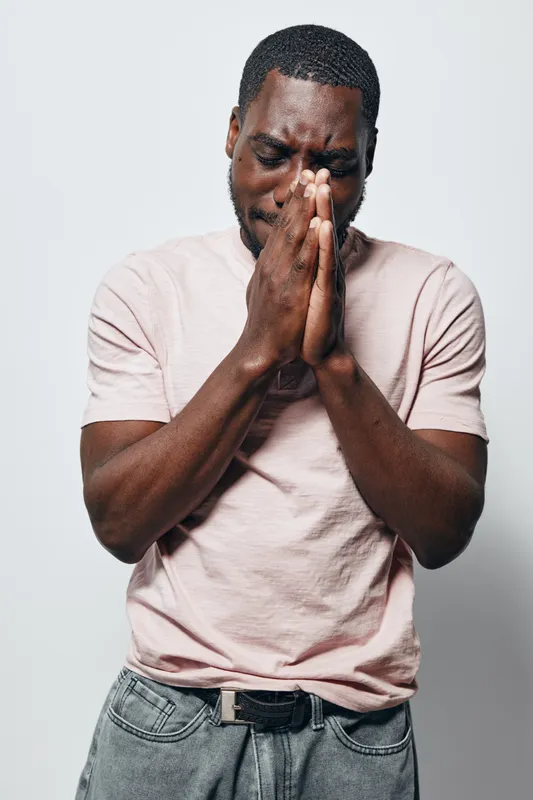 African American man with dark skin wearing light pink t shirt and jeans standing with hands clasped in prayer or deep thought against white background.