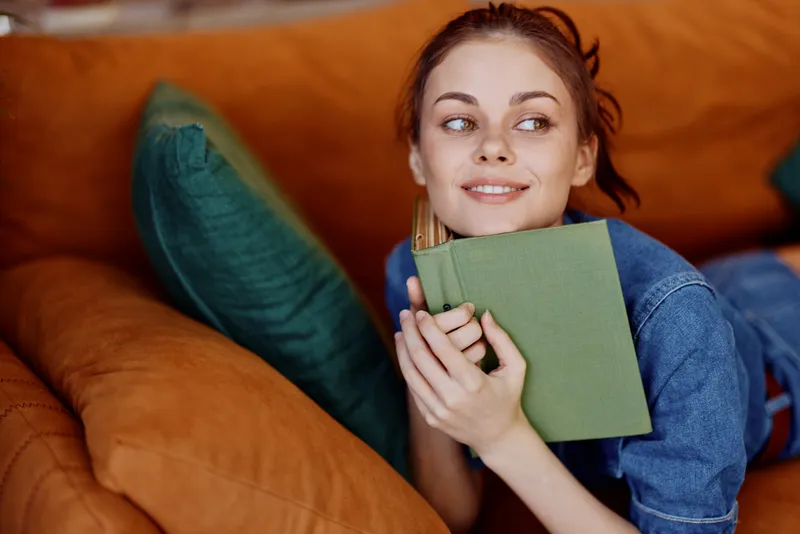 Счастливая женщина лежит на диване с книгой в руке и улыбкой на лице