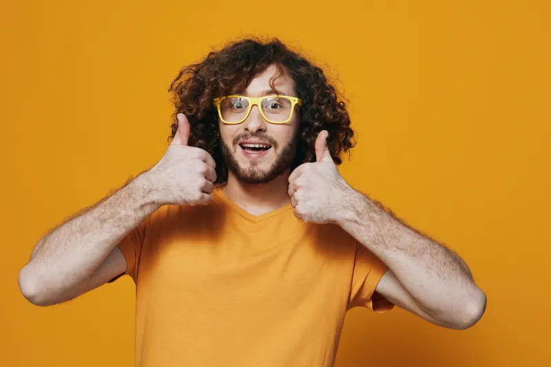 Happy young man with curly hair wearing yellow glasses and an orange t shirt, giving a thumbs up against a bright yellow background, conveying positivity and joy