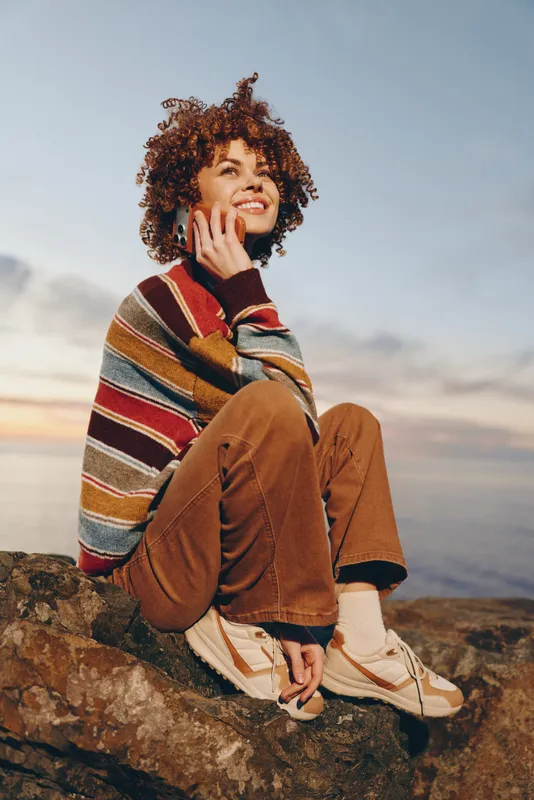 Woman outdoors smile sunset mountains lifestyle striped sweater backpack curly hair, laughing softly holding hand near face, sitting on rock at evening, warm light, casual travel outfit, natural