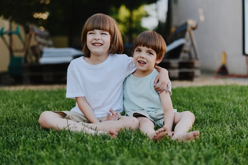 Two joyful boys sitting together on grass, smiling brightly in a sunny outdoor setting. Their camaraderie captures the essence of childhood friendship and happiness.