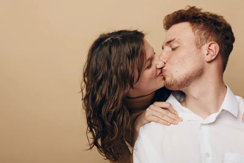 Young couple kissing tenderly with closed eyes on beige background. Intimate moment shows love, affection, and connection between man and woman in casual white shirt and natural hair.