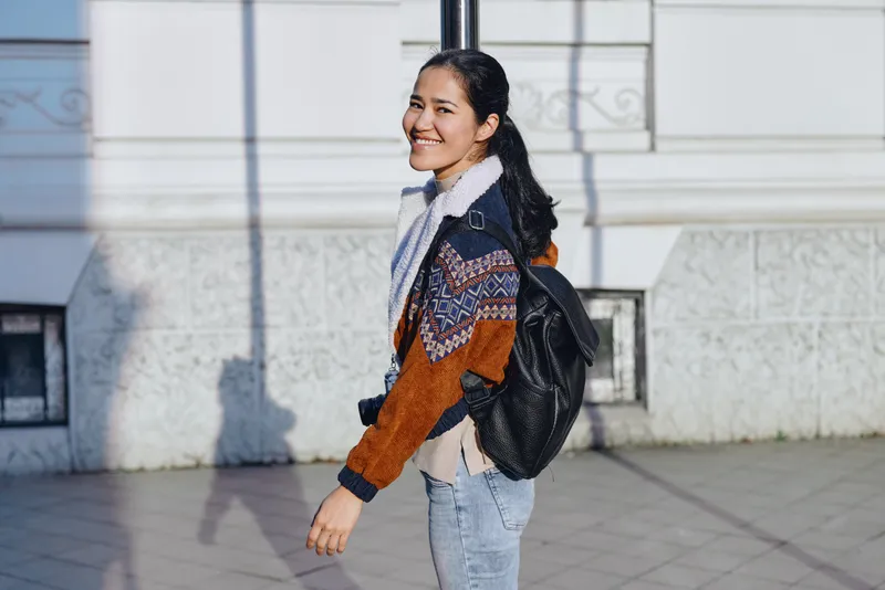 Backpack, street, walking, smile, jacket, casual, urban fashion, travel, outdoor scene, denim and sweater, sunlight on pavement, capturing a relaxed, friendly mood during a city stroll