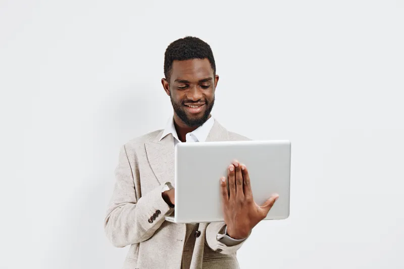 Confident young man with dark skin dressed in a light suit holding a laptop while smiling and looking at the screen. Studio portrait on plain white background, conveying technology, professionalism, a