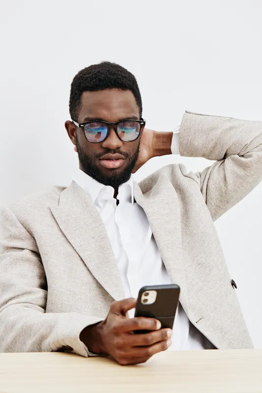 Stylish young man wearing glasses and beige blazer using smartphone while sitting at table against plain white background. People lifestyle concept, modern, casual, social media, communication, urban.