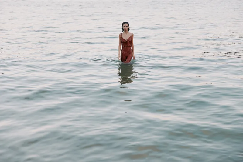 A woman stands waist-deep in calm sea water wearing a red dress; tranquil scene, soft light, reflective surface, subtle waves, and a serene mood.