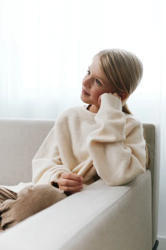 Young girl sits on a light sofa wearing a cozy cream sweater, gazing thoughtfully to the side, conveying calm, warmth, and gentle introspection in a bright living space.