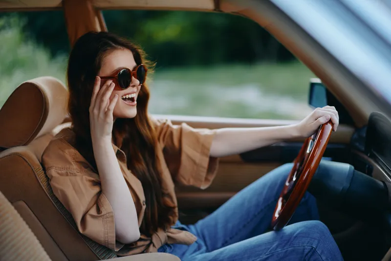 Happy young woman in sunglasses enjoying a road trip in a stylish car, wearing casual clothes, with a joyful expression against a green background.