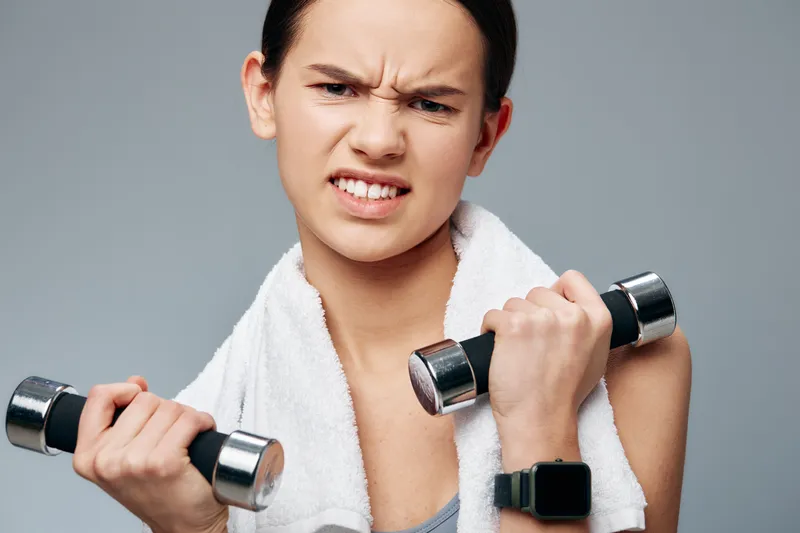 Fitness and sport themed image of a focused woman lifting dumbbells, with a towel around her neck and a smartwatch, during a gym workout and training session.