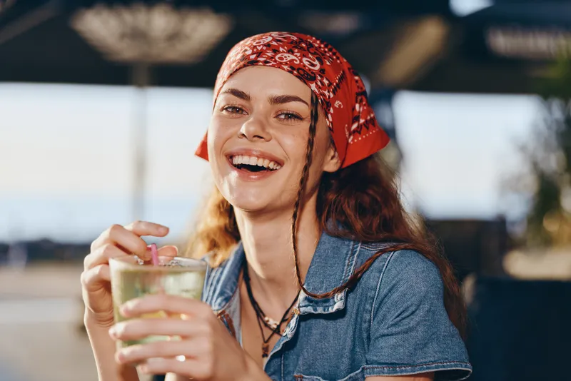 Lifestyle woman with modern boho-western denim street style smiling joyfully outdoors holding a drink in film color warm tones and relaxed mood