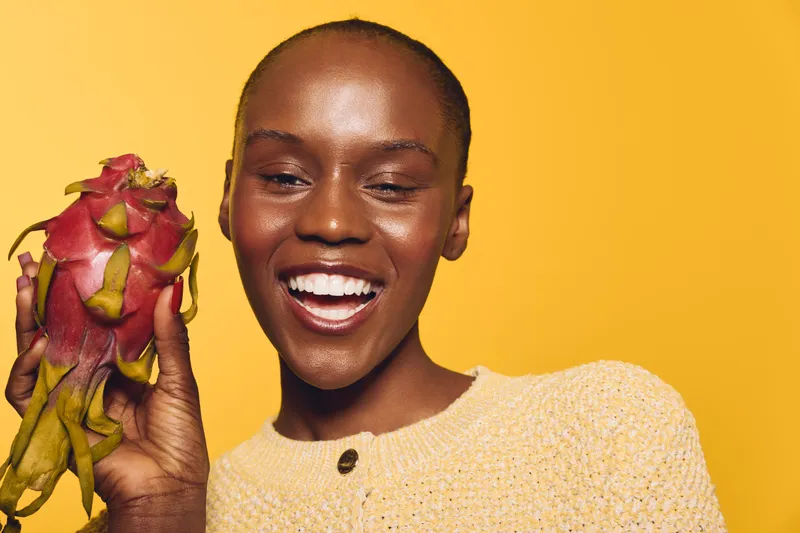 Happy African woman with dark skin holding dragon fruit, smiling with teeth, wearing yellow sweater. Close-up on yellow background. Concept of healthy lifestyle, fruit, and natural beauty.