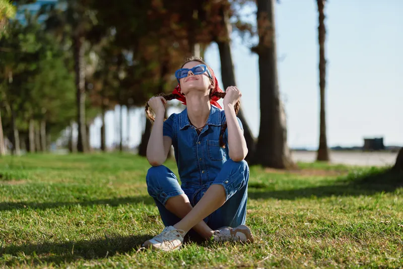 Lifestyle woman in modern boho-western denim street style sitting on grass with sunglasses enjoying sunny day, film color mood and relaxed outdoor vibe.