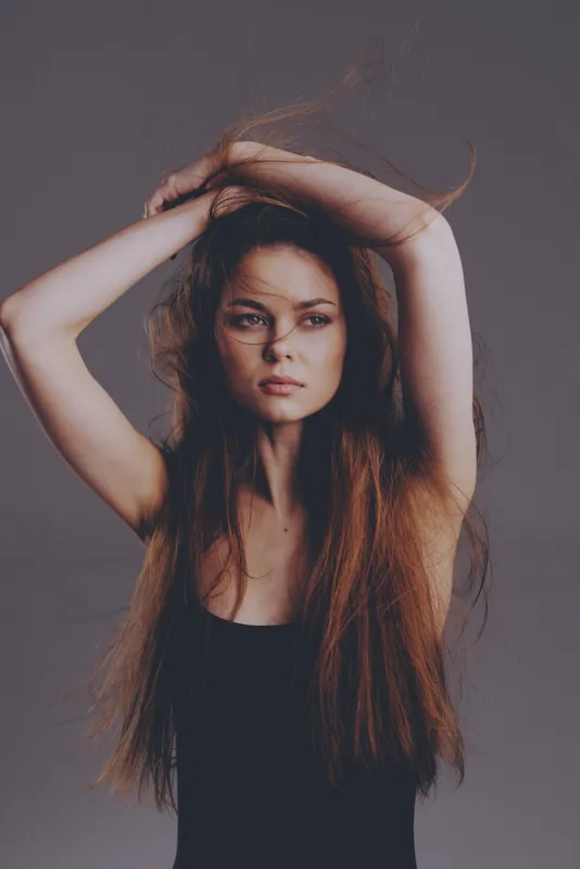 Portrait of a serious woman with long brown hair and expressive eyes wearing a black top in a minimalistic studio setting.