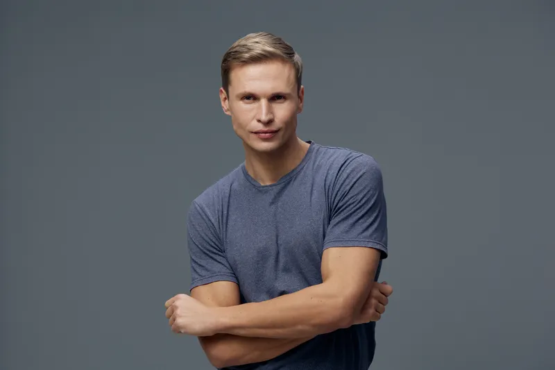 Confident young man with short hair wearing casual blue t-shirt looking at camera in studio setting isolated on plain gray background. Studio portrait. People lifestyle concept.