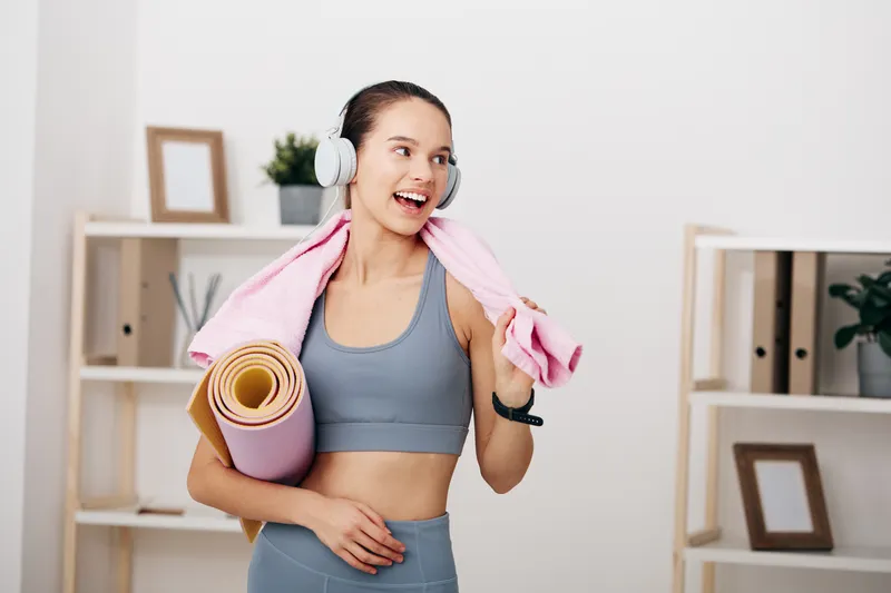 Fitness woman in workout attire with headphones, holding a yoga mat and towel in a home gym, smiling after exercise and ready for the next set.