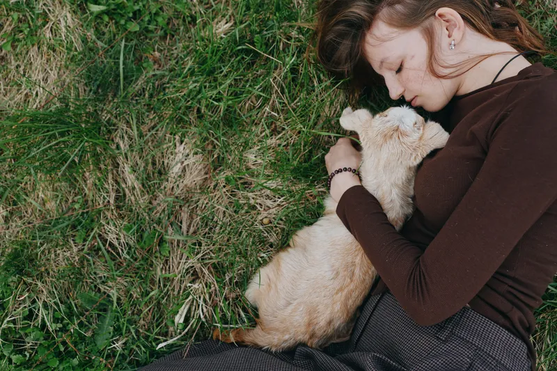 A woman lies on grass, cuddling a light furred animal in a peaceful outdoor moment. A candid lifestyle shot conveying comfort, affection, and connection with a pet.