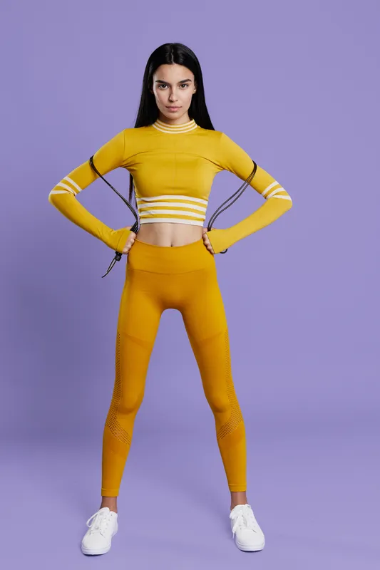 Woman sport fitness dressed in yellow activewear stands confidently with hands on hips, showcasing strength and determination against a soft purple background.