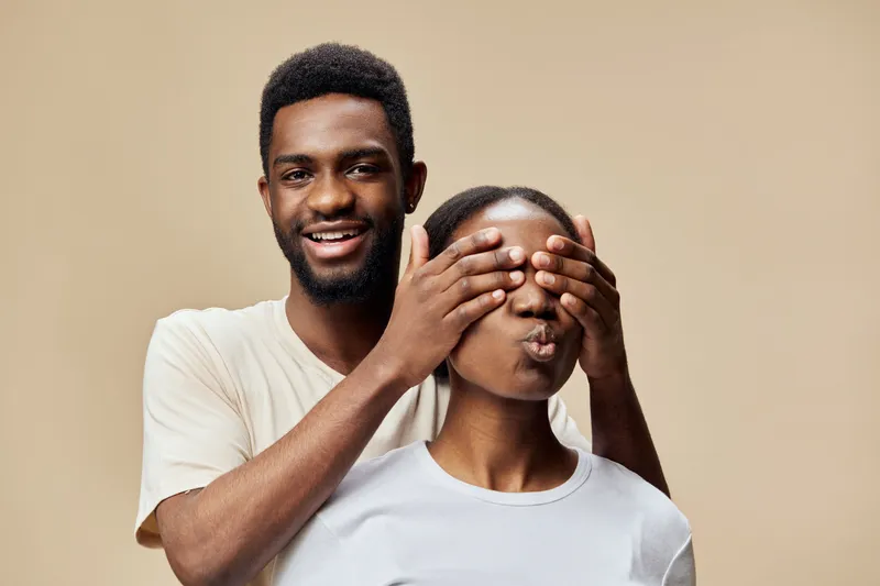Happy couple playfully engaging in a fun moment together, with the man playfully covering the woman s eyes against a soft beige background