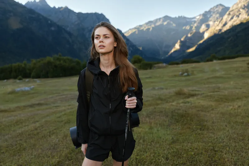 A confident woman explorer stands in a rugged mountain valley, wearing black hiking gear and a backpack, gazing ahead as sunlight highlights the dramatic scenery.