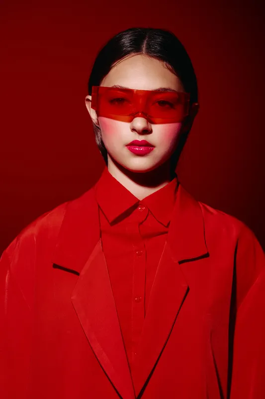 Young woman in red coat and giant sunglasses poses with slick hair and striking lip gloss on red background. Photo concept of beauty creative fashion and bold style.