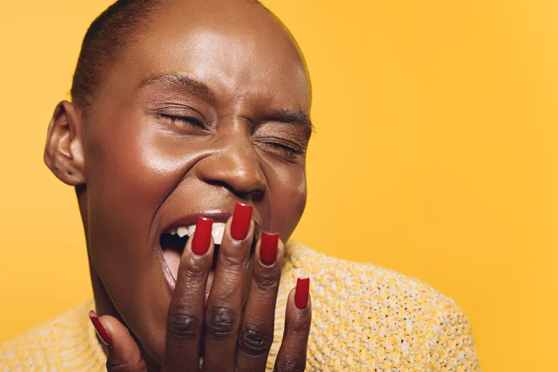 Close-up of joyful African woman with dark skin laughing with hand near mouth. Yellow background and textured yellow sweater create warm, happy, and positive mood concept of joy and emotion.