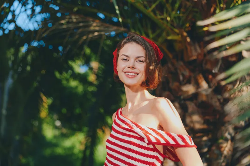 An upbeat woman poses outdoors in a sunlit garden, wearing a red and white striped off-shoulder top. She smiles warmly, projecting confidence and charm amid lush greenery and bright natural light.