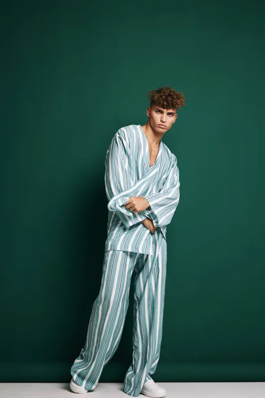 Young man with curly hair wearing striped pajamas and white sneakers standing against green background. Casual pose with arms crossed and relaxed expression in studio setting.