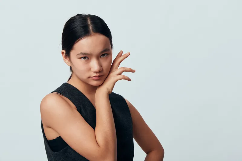 Young woman posing with hand near face and looking at camera, wearing sleeveless black top in simple studio against light gray background. Portrait of confident female beauty model.
