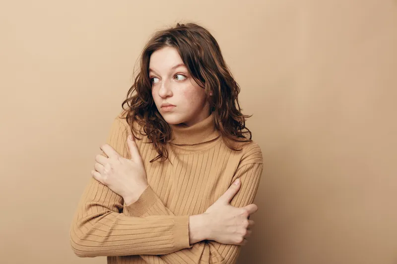 Young woman in beige sweater looks sideways with crossed arms, expressing discomfort or cold. Neutral background highlights her thoughtful and uneasy mood in casual clothing.