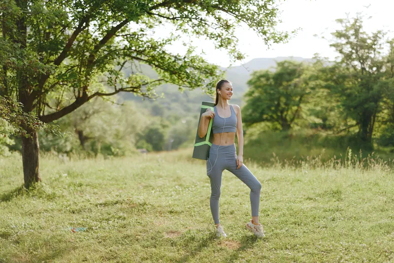 Fit woman in athletic wear holding a yoga mat, enjoying nature in a fitness focused setting, embodying health and vitality during sunny outdoor training