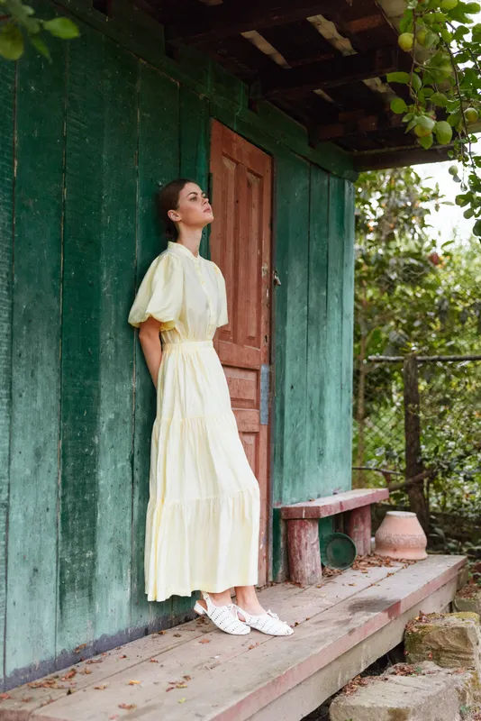A woman in a pale yellow dress stands on a wooden porch beside a green wall, enjoying the outdoors with a calm, contemplative pose in natural light.