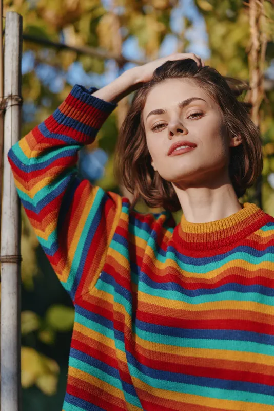 Woman portrait colorful sweater outdoor autumn, young casual model with confident gaze, hand in hair and natural sunlight highlighting knitwear and warm leaves backdrop