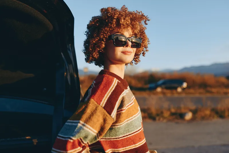 Woman lifestyle, curly hair and sunglasses, standing by a parked car on an open road. Outdoor travel moment at golden hour, striped sweater, mountains and blue sky, casual.