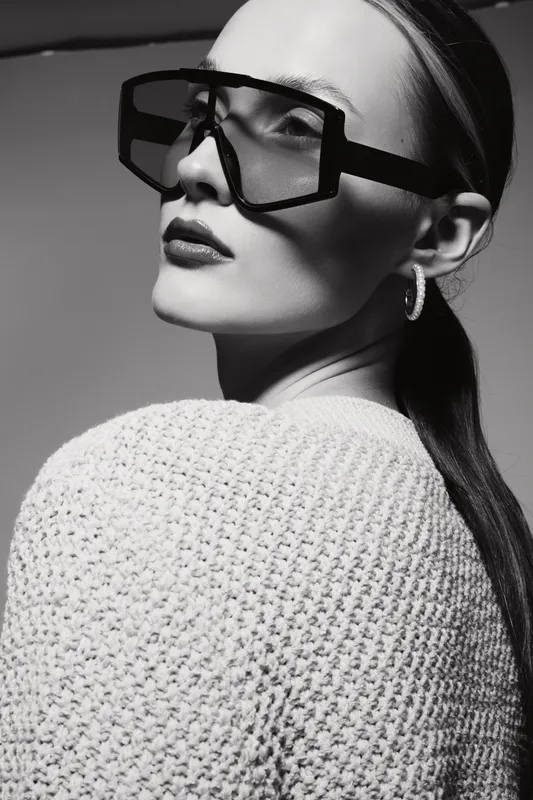 Portrait of a fair skinned Caucasian woman wearing large geometric sunglasses and hoop earrings in soft lighting on grey background.