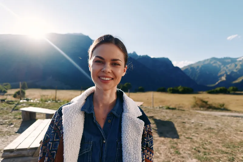 Woman smile portrait in mountains outdoors with sunlight and nature landscape, young happy traveler in denim jacket by picnic bench, candid headshot during golden hour adventure.