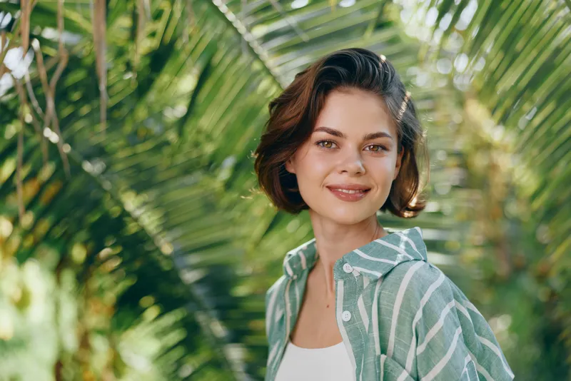 Portrait of a smiling woman outdoors among tropical greenery, wearing a striped green shirt and white top, bathed in natural light.