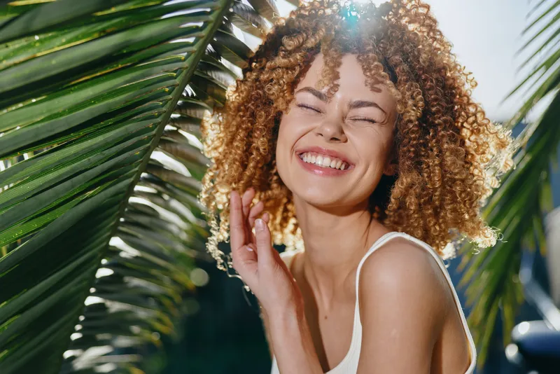 Smiling woman with curly hair enjoys sunny tropical outdoors, surrounded by green palm leaves, radiating happiness and natural beauty in warm daylight.