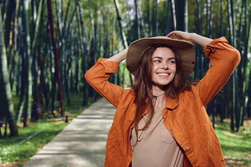 Woman smile hat bamboo forest path jacket outdoors portrait of a happy young traveler wearing a straw hat and orange jacket on a sunlit walkway through tall green bamboo trees.