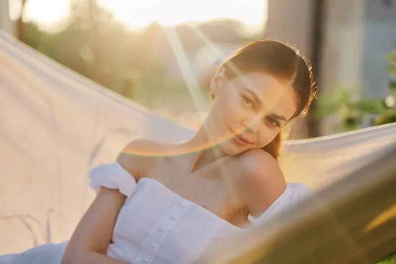 Young woman relaxing in a hammock, wearing a white dress, enjoying sunset vibes with a serene expression, surrounded by nature in soft pastel tones