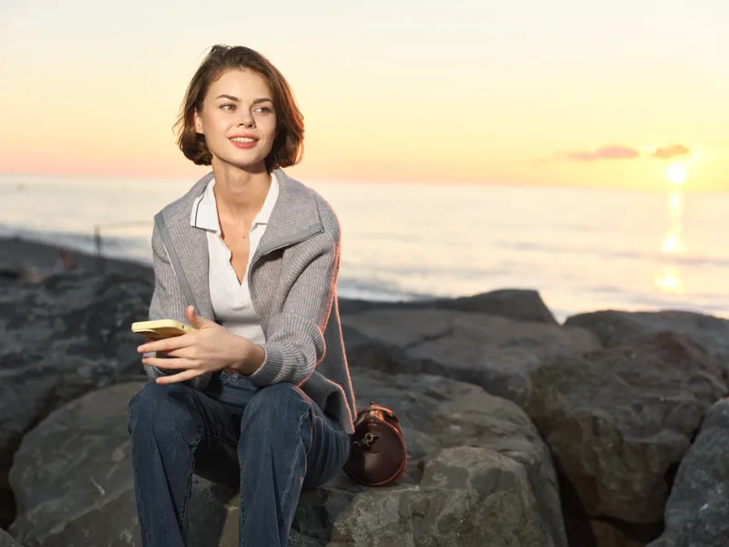 A smiling woman sits on rocky shoreline at sunset, using a mobile phone. She wears a light blazer and jeans, conveying calm focus and a relaxed, productive mood.