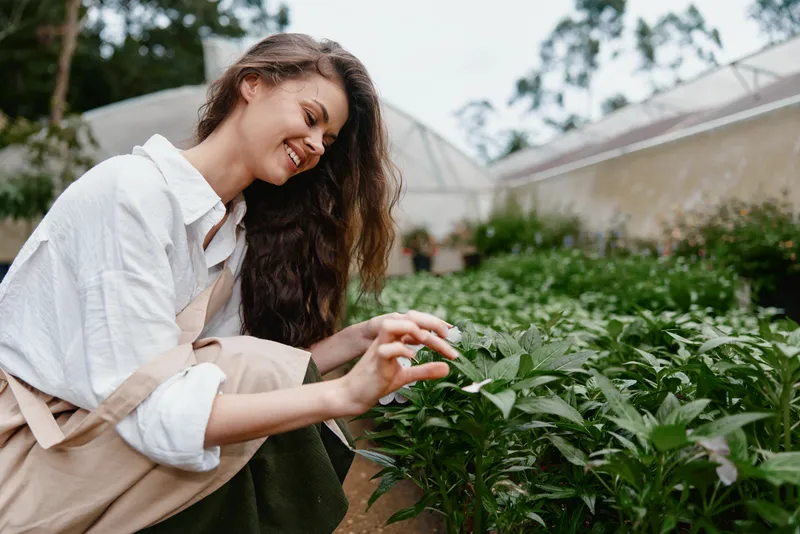 Young woman with long hair smiling while tending to green plants in a garden, showcasing sustainable gardening practices and a connection to nature