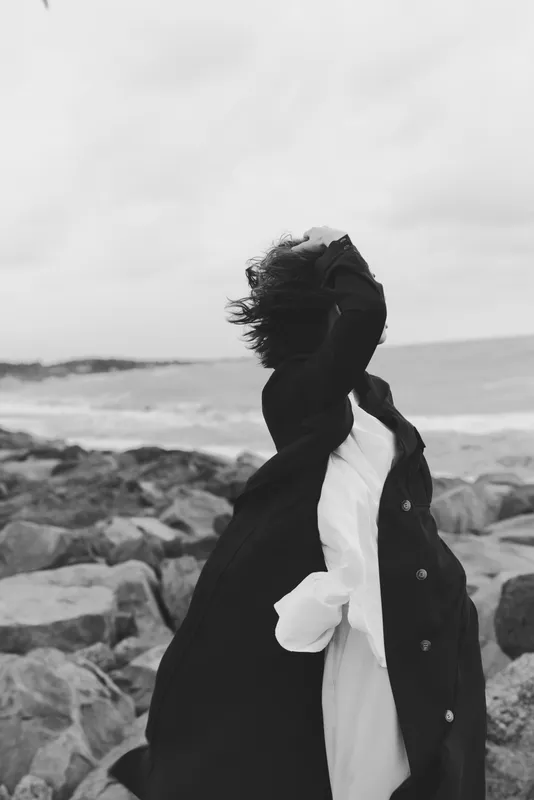 Wind sweeps along a rocky coast as a woman in a flowing coat shields her face, creating a dramatic silhouette against the sea and overcast sky, conveying mood, movement, and rugged coastal atmosphere.