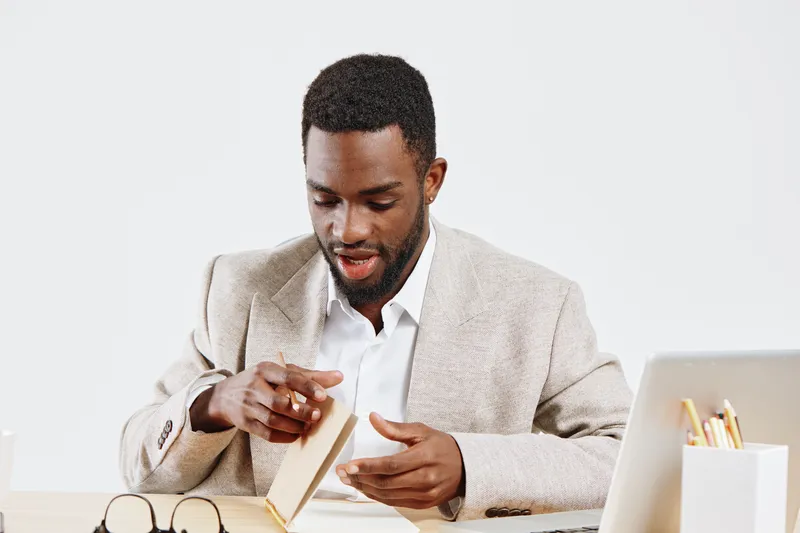 Businessman in beige blazer reading a notebook at his desk, surrounded by office supplies, laptop, and glasses, focusing on his work, in a professional modern office setting, concept: workplace produc