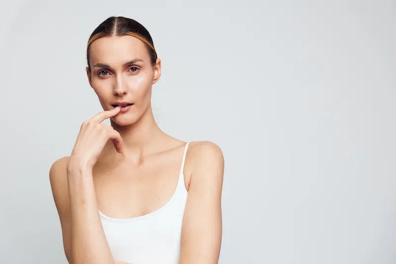 Young woman with natural makeup posing with finger near lips, wearing white tank top, neutral expression, clean background, soft lighting, minimalistic style portrait.