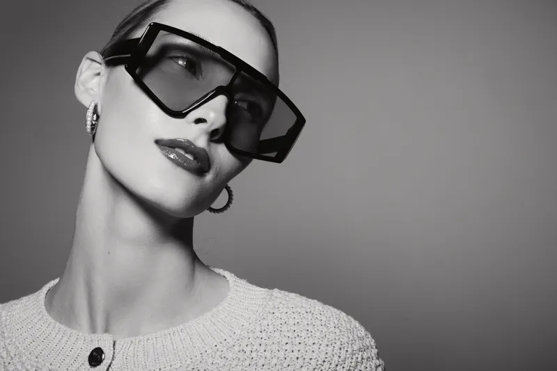 Portrait of a young white woman with fair skin wearing oversized black sunglasses and hoop earrings posing against a plain background.