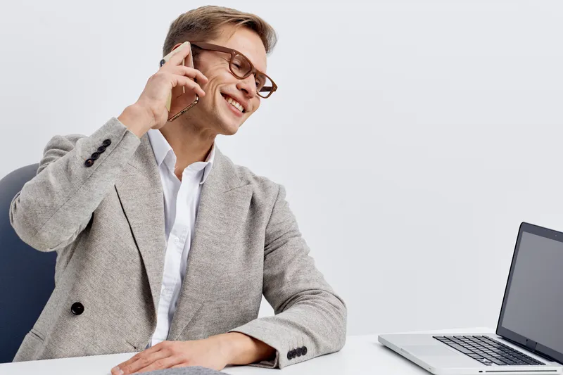 Smiling young caucasian man in grey blazer talking on smartphone sit at white table with laptop work remotely digital nomad concept