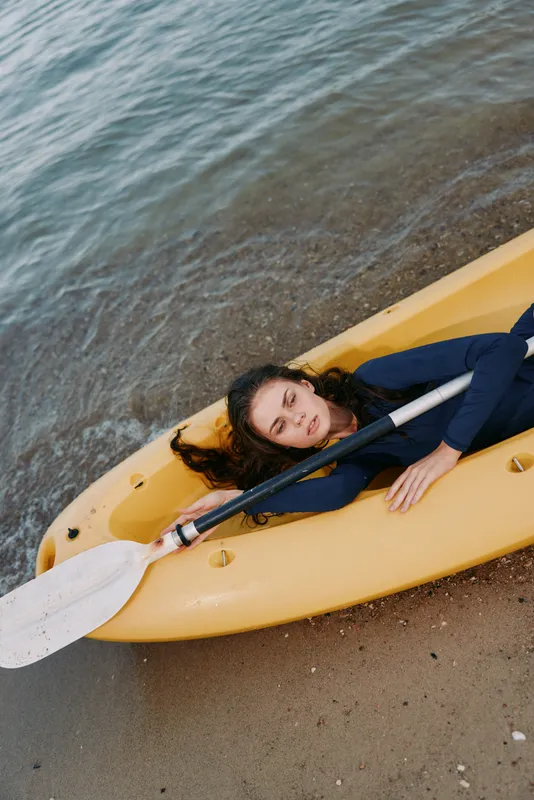 A person lies inside a yellow kayak resting on a sandy shore, calm water nearby, relaxed pose and quiet moment of outdoor exploration and leisure.