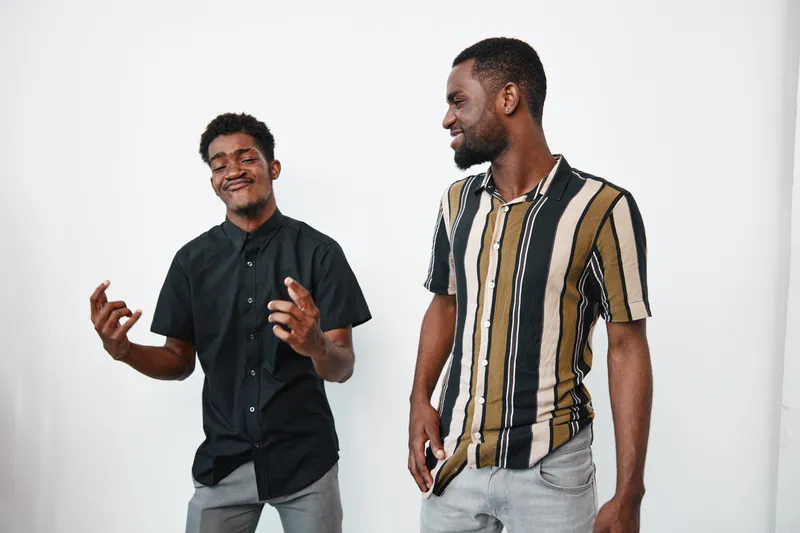 Two young African American men with dark skin standing and smiling together in casual clothes on white background. One wears black shirt, other striped shirt with short sleeves.